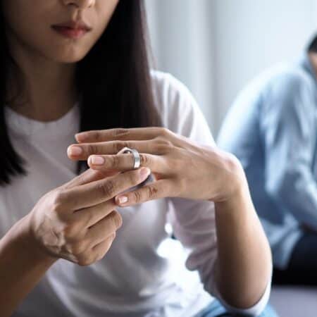 woman-taking-off-wedding-band-as-frustrated-husband-sits-in-background